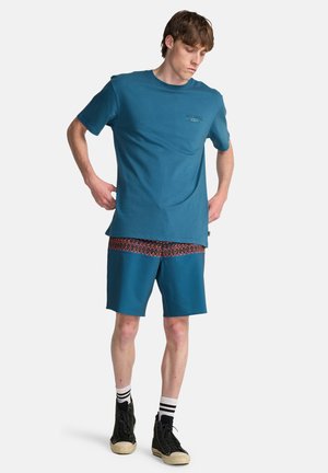 Niebieski bawełniany t-shirt z małym logo, w połączeniu z niebieskimi szortami z czerwonym wzorzystym paskiem w pasie. Czarne trampki na wysokiej cholewce z białymi paskami.