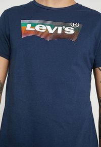 Mörkblå bomullströja med en flerfärgad Levi's-logotyp och horisontella ränder över bröstet. Korta ärmar, rundad halsringning.