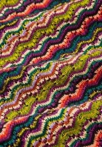 Bunt gestrickter Stoff mit einem wellenförmigen Zickzack-Muster in Grün, Lila, Orange und Creme, versehen mit strukturierten Stichen und subtilen Schimmer.