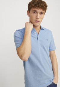 Hellblaues Poloshirt mit Kragen, drei Knöpfen und kleinem Logo auf der Brust. Kurzarm und strukturierter Stoff.