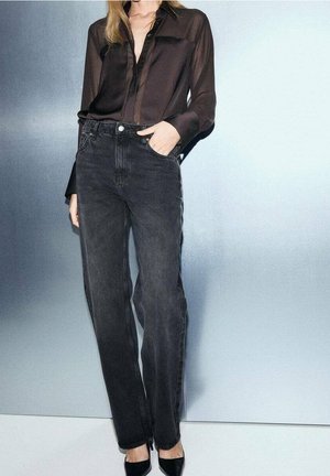 Mujer con blusa negra transparente, jeans negros de cintura alta y tacones negros puntiagudos sobre un fondo gris claro.