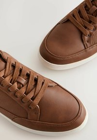 Een paar bruin leren vetersneakers met witte zolen tegen een lichte achtergrond, met gedetailleerde stiksels en gestructureerde zijpanelen.
