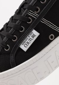 Detailní záběr na černou plátěnou tenisku s černými tkaničkami, bílým dvojitým prošíváním, textilní štítkem "Versace Jeans Couture" a bílou embosovanou podrážkou.