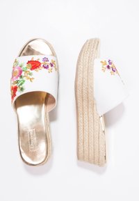 Sandales slides blanches avec motif floral brodé, semelle intérieure métallique dorée, et semelle plateforme en jute. Dotées d’une semelle épaisse et texturée ainsi que d’une lanière plate.