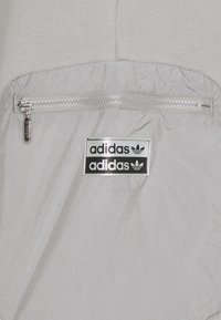 Grey athletic jacket with a zip pocket, made of textured fabric. Features two black Adidas logo labels and silver zipper hardware.