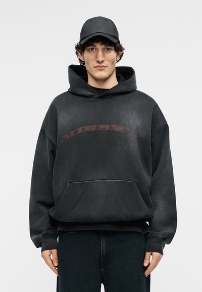 Νεαρός άνδρας με ξεθωριασμένο μαύρο φούτερ με μπροστινή τσέπη και μαύρο καπέλο μπέιζμπολ, στέκεται μπροστά σε απλό φωτεινό φόντο.