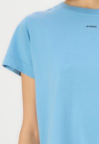 Ανοιχτό μπλε βαμβακερό μπλουζάκι με κοντά μανίκια, στρογγυλή λαιμόκοψη και ένα μικρό μαύρο λογότυπο "PINKO" κοντά στο γιακά. Λείος υφαντός.
