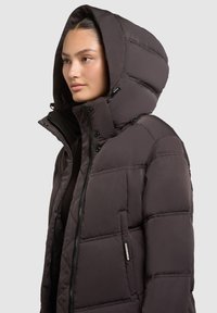 Chaqueta de plumas marrón oscura con un cuello alto y una gran capucha, que cuenta con un cierre de cremallera y detalles de botón, textura suave y diseño acolchado.