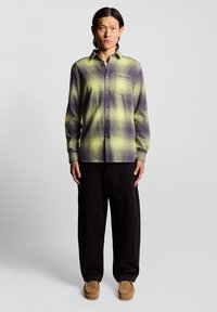 Kariertes Hemd in grünen und lila Tönen, mit Knöpfen und Kragen; getragen mit schwarzen Hosen und braunen Schuhen. Glattes Gewebe mit einem Schottenmuster.