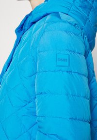 Ljusblå vadderad jacka med huva och BOSS-logotyp på övre ärm, bärs av en person som visas från axel till bröst.