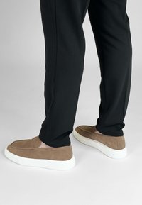 Bruine suède instapschoenen met een witte rubberen zool, gezien van achter, gecombineerd met zwarte taps toelopende broek. Geen zichtbare logo's of patronen.