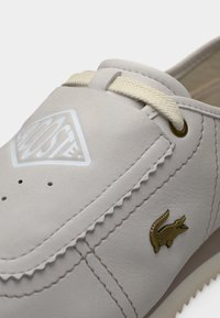Chaussure en cuir gris clair avec détails en lacets, design cousu, perforations sur l'avant, et logo crocodile doré sur le côté.