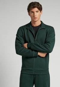Sudadera de cremallera en verde oscuro, hecha de un tejido suave, con un cuello alto y puños elásticos, llevada con una camiseta negra debajo.