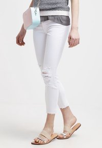 Jeans skinnies blancos desgastados con dobladillos deshilachados, combinados con una blusa gris y un bolso bandolera azul claro. Sandalias planas plateadas y beiges.