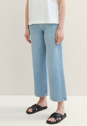 Person trägt hellblaue cropped Wide-Leg-Jeans mit weißem Oberteil und schwarzen, aus Leder gefertigten Crisscross-Slippern.