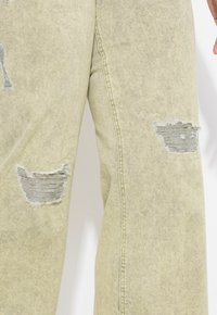 Pantalons en denim vert clair avec des zones effilochées présentant des bords effilés, une coupe décontractée et des coutures visibles le long des coutures.