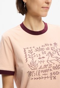 Φωτεινό ροζ κοντομάνικο μπλουζάκι με μπορντό ριμπ γιακά. Διαθέτει περίπλοκη μπορντό κεντητική διακόσμηση με φυτά και ζώα στο μπροστινό μέρ ος.