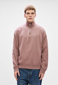 Svetle růžová mikina s vysokým límcem a detailním zipem na půl, vyrobená z měkké tkaniny. Má volný střih a žebrované manžety.