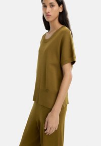 Oliwkowy sweter z krótkim rękawem i okrągłym dekoltem. Posiada żebrowane wykończenie na dole oraz mały detal z logo.