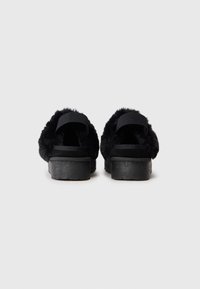 Schwarze Plattformsandalen mit flauschiger schwarzer Felltextur, ausgestattet mit einem gepolsterten Riemen und einer festen schwarzen Gummisohle. Die Rückansicht zeigt die Riemenposition.