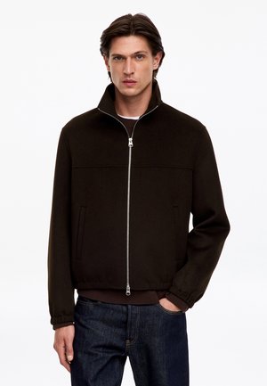Jeune homme aux cheveux bruns portant une veste noire zippée et un jean bleu foncé, debout avec une main dans la poche devant un fond blanc.