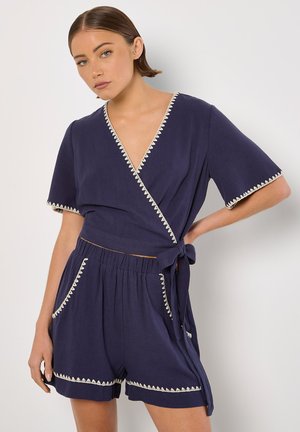 Top avvolgente blu navy con maniche corte, caratterizzato da un bordo a zigzag bianco lungo il collo e le maniche. Abbinato a pantaloni corti coordinati con tasche.