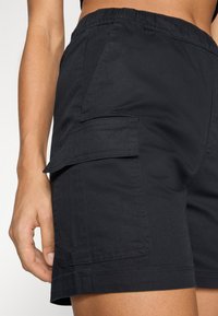 Svarta cargoshorts gjorda av mjukt tyg, med sidoficka, elastiskt midjeband och sömmar. Närbild på detaljerna för midjebandet och fickan.