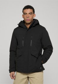 Zwarte, hooded jas met ritszakken op de borst, een gladde textuur en een getailleerd ontwerp, gecombineerd met lichtbruine broek.