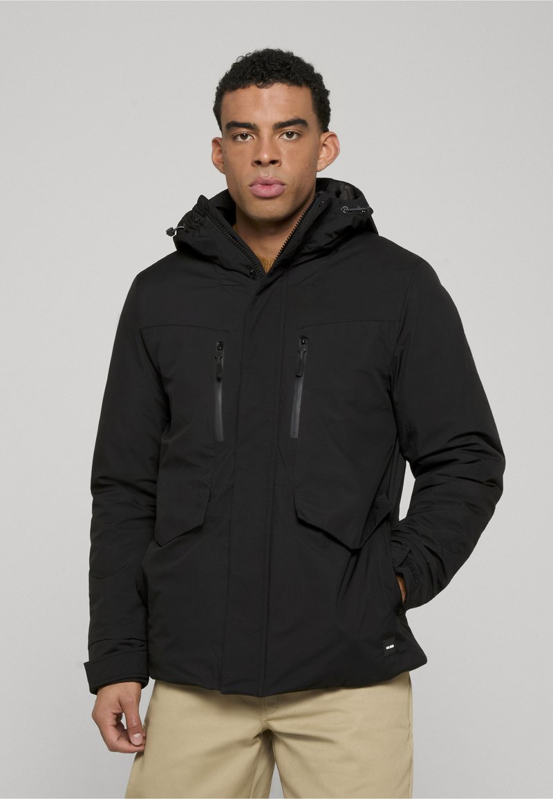 Zwarte, hooded jas met ritszakken op de borst, een gladde textuur en een getailleerd ontwerp, gecombineerd met lichtbruine broek.