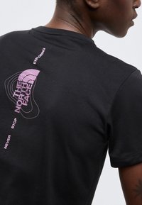 Άτομο που φοράει μαύρο μπλουζάκι The North Face με μοβ λογότυπο και κείμενο "Ποτέ μην σταματάς να εξερευνάς" στο πάνω μέρος της πλάτης.