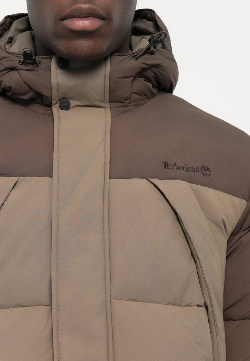 Chaqueta de plumón marrón y marrón claro con cremallera en la parte delantera, capucha y el logo de Timberland en el pecho. La tela es suave y presenta un diseño acolchado.