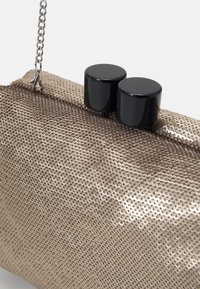 Złota metaliczna kopertówka o cekinowej teksturze, z łańcuszkowym paskiem i dwoma czarnymi cylindrycznymi zapięciami na górze do zamknięcia.
