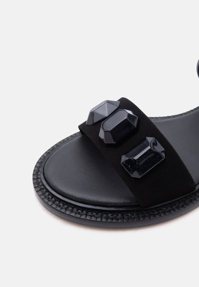 Μαύρο σανδάλι slide με τρεις μεγάλες πολύπλευρες μαύρες πέτρες ως διακόσμηση στον φαρδύ ιμάντα πάνω από τα δάχτυλα, ορατή υφή στην άκρη της σόλας.