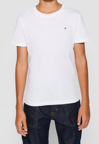 Camisa de algodón blanca con cuello redondo, mangas cortas y un pequeño logo en el lado izquierdo del pecho. Combinada con jeans de mezclilla oscura.