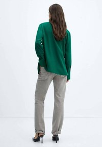 Chemise verte oversize à manches longues associée à un pantalon taille haute gris clair et des chaussures à talons noires. L'arrière-plan est blanc.