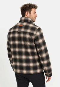 Zwart en beige geblokte flanellen jack met een kraag, knoopsluiting en leren accenten op de rug, ontworpen voor warmte en duurzaamheid.