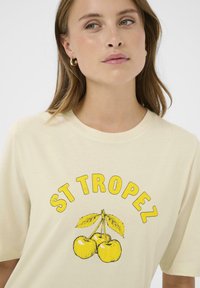 Femme portant un t-shirt crème avec des cerises jaunes et le texte "ST TROPEZ" en lettres jaunes grasses sur la poitrine.