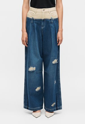 Jean bleu large avec une ceinture haute beige, des patches usés sur les jambes, porté avec des talons blancs à bout pointu et un haut noir.