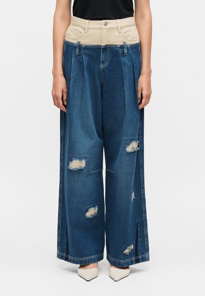 Jean bleu large avec une ceinture haute beige, des patches usés sur les jambes, porté avec des talons blancs à bout pointu et un haut noir.
