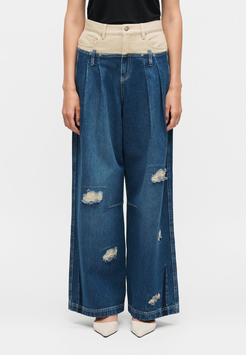 Jean bleu large avec une ceinture haute beige, des patches usés sur les jambes, porté avec des talons blancs à bout pointu et un haut noir.