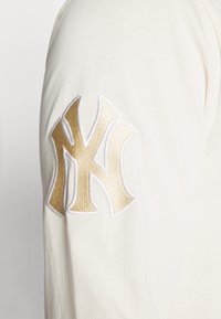 Cream-colored long-sleeve shirt featuring a gold and white embroidered New York Yankees logo on the left sleeve. Soft, smooth fabric texture.
