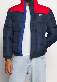 Man i marinblå och röd pufferjacka över vit skjorta och ljusblå jeans, med handen i jackfickan och jackan delvis uppknäppt.