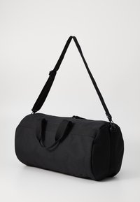 Bolsa de deporte negra hecha de una tela resistente, con cierre de cremallera, doble asa y una correa para el hombro ajustable. Textura suave, forma cilíndrica.