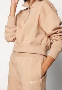 Sudadera de color beige claro con cremallera hasta un cuarto, puños y cinturilla acanalados; cuenta con un pequeño logo blanco de Nike en el pecho y pantalones joggers a juego.