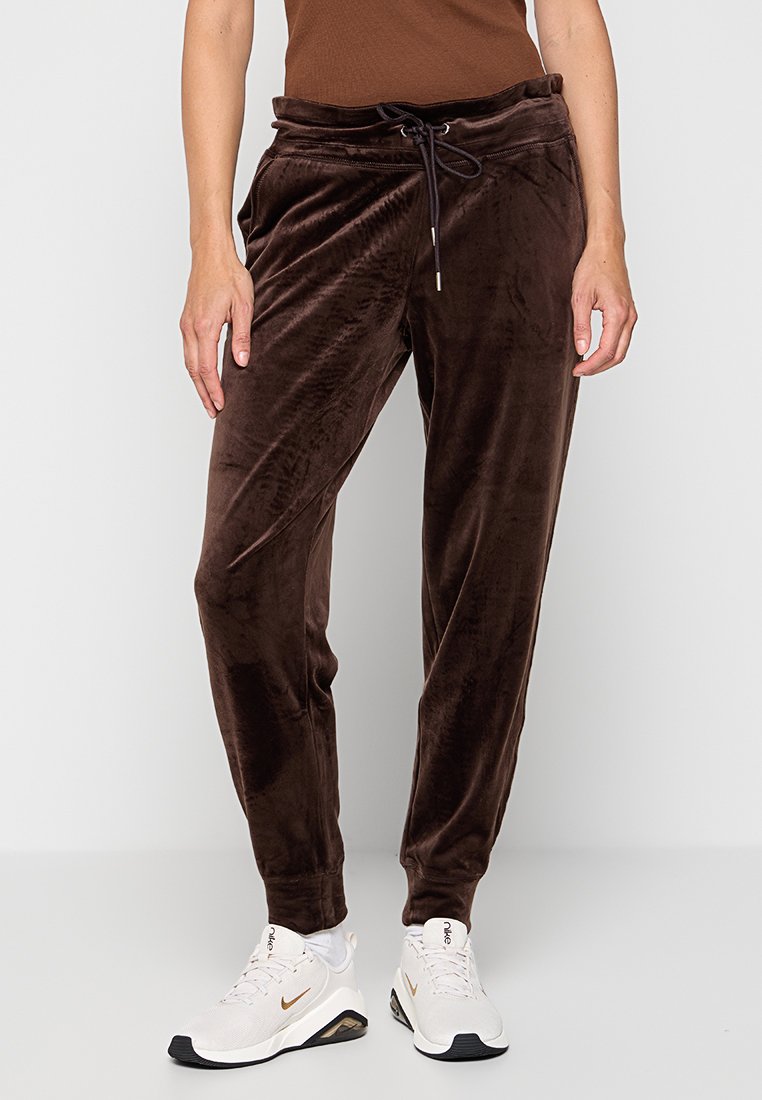 Brązowe dresowe spodnie z weluru z elastycznym pasem ze sznurkiem, bocznymi kieszeniami i zwężanym krojem, w połączeniu z białymi sneakersami. Widoczna miękka tekstura i połysk.