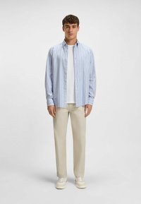 Niebiesko-biała w paski koszula zapinana na guziki z długim rękawem, zestawiona z beżowymi spodniami i białymi sneakersami, noszona przez stojącego mężczyznę.