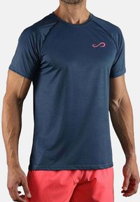 Hombre vestido con una camiseta deportiva de manga corta azul marino con un pequeño logo rosa y pantalones cortos rojos coral, de pie contra un fondo liso.