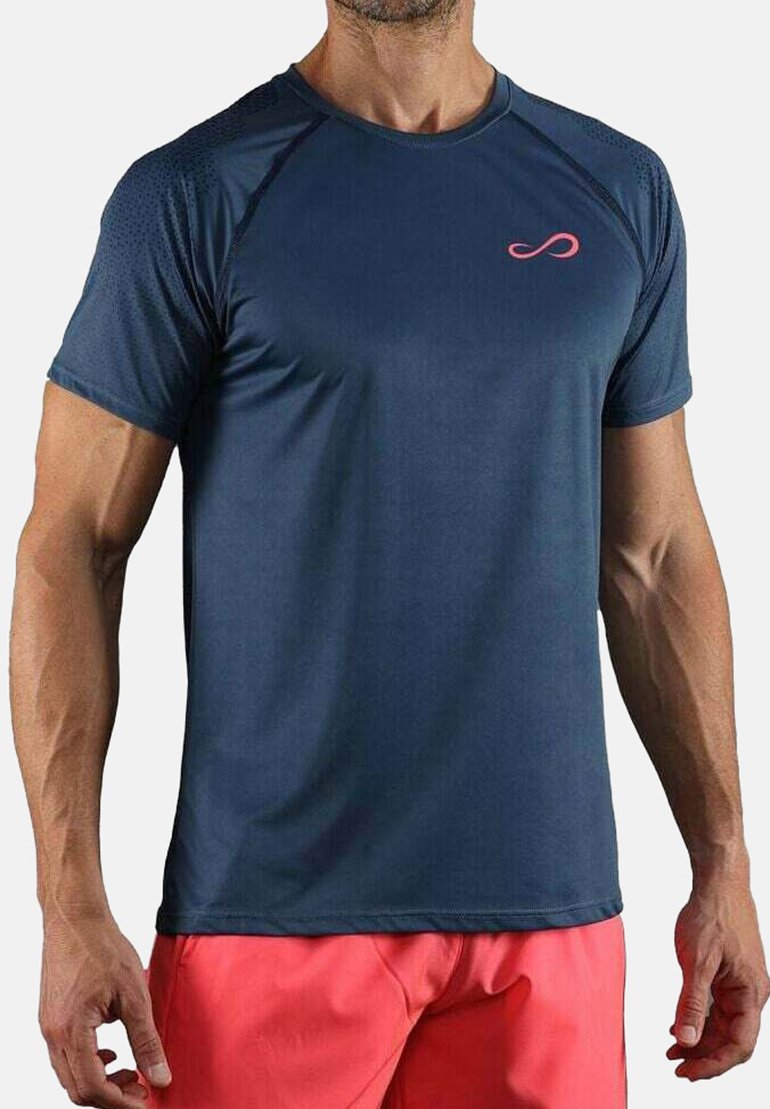 Hombre vestido con una camiseta deportiva de manga corta azul marino con un pequeño logo rosa y pantalones cortos rojos coral, de pie contra un fondo liso.