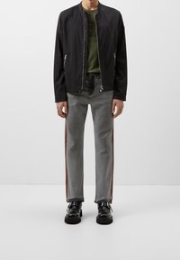 Chaqueta bomber negra, camiseta de color verde olivo, jeans grises con franjas laterales naranjas, zapatos negros con acentos metálicos. Tejido suave y ajuste entallado.