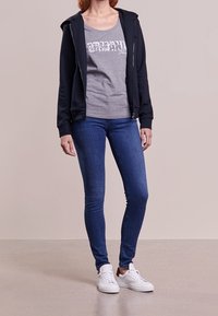 Marinblå zip-up hoodie över en grå tanktop med en glittrande grafik, tillsammans med blå smala jeans och vita sneakers.